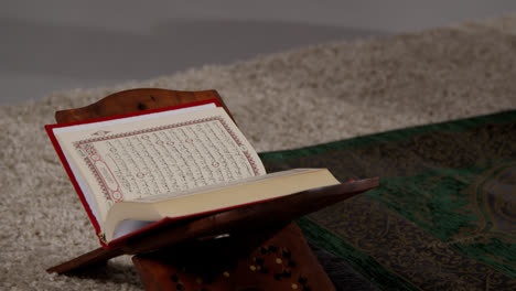 Close-Up-Of-Open-Copy-Of-The-Quran-On-Stand-Next-To-Prayer-Mat-In-Muslim-Home-1